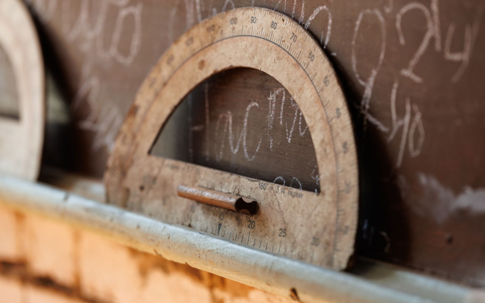 An old wooden protractor rests on a chalkboard tray covered in faded chalk writing