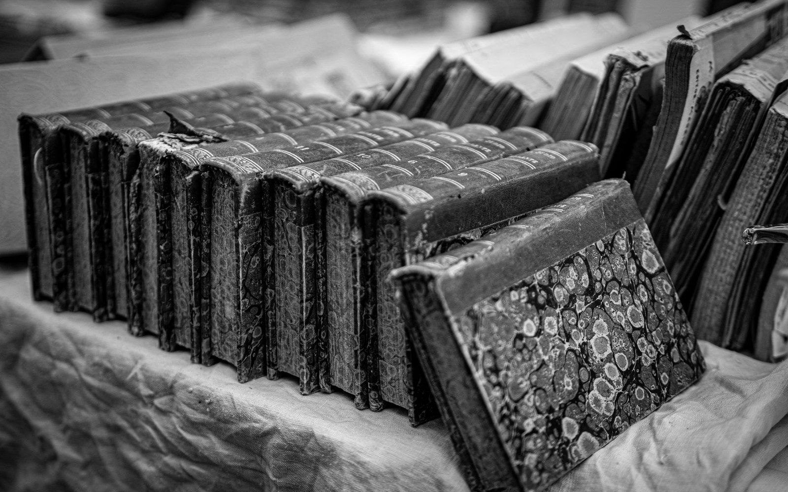 Rows of worn antique books with marbled covers lean together on a cloth-covered table