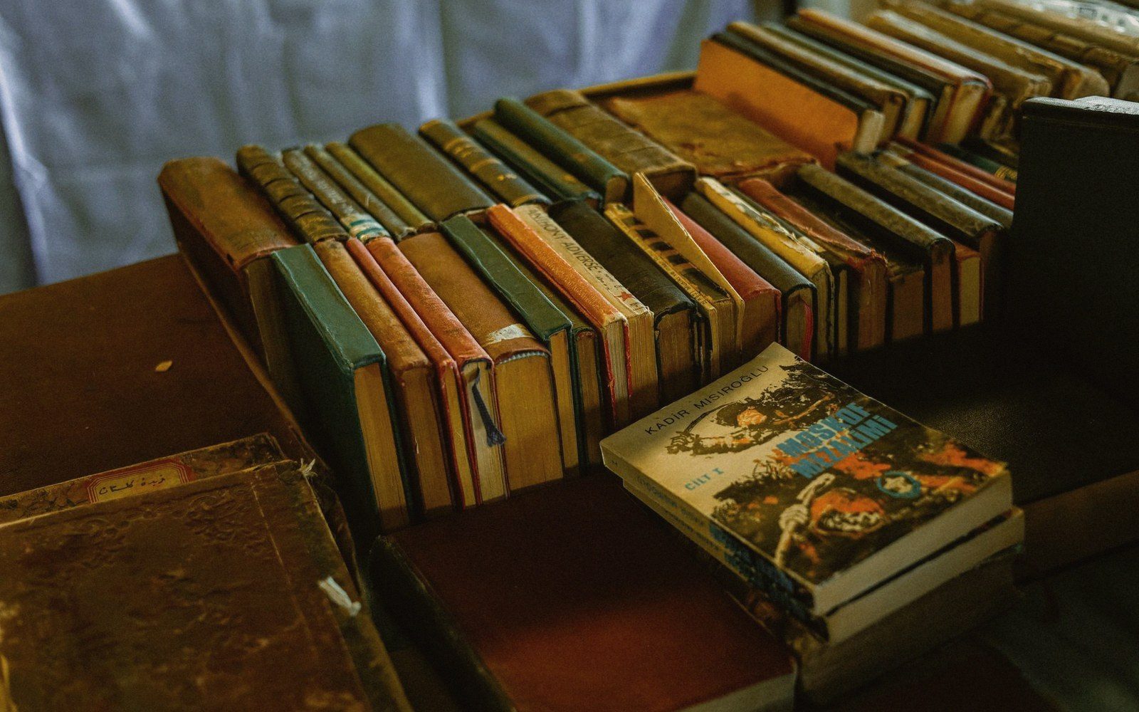 Rows of worn hardcover books stacked on a wooden table beside a paperback novel